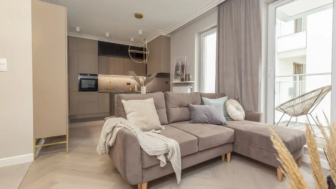 Nowoczesny salon z aneksem kuchennym. Dominują barwy monochromatyczne przykłady: beże, szarości, brązy. Miękka sofa, jasne drewno.