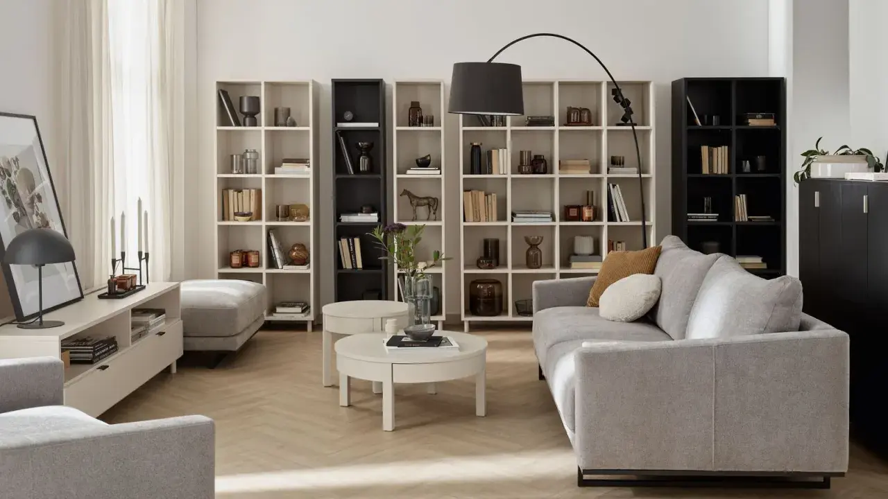 Nowoczesny salon beżowo szary z szarą kanapą, białymi regałami i czarną szafką.