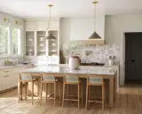 Jasna kuchnia w stylu modern classic z wyspą i czterema hokerami. Jasne meble, marmurowy blat, złote detale.