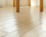 Jasna podłoga z drewna, gdzie widać, że są za ciasno ułożone panele. Światło odbija się od gładkiej powierzchni.