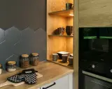 Nowoczesne półki w kuchni aranżacje: drewniane, otwarte, z podświetleniem, pełne kubków i kieliszków. Obok słoiki z produktami i rękawica kuchenna.