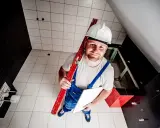 Budowlaniec w kasku i ogrodniczkach trzyma poziomicę i płytkę. Pokazuje, jak skuć płytki bez kurzu.