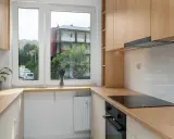 Jasna, nowoczesna mała kuchnia z oknem na środku, przez które widać budynki i drzewa. Drewniane blaty i szafki tworzą przytulną atmosferę.
