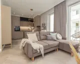 Nowoczesny salon z aneksem kuchennym. Dominują barwy monochromatyczne przykłady: beże, szarości, brązy. Miękka sofa, jasne drewno.