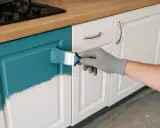 Ręka w rękawiczce maluje pędzlem turkusową farbą fronty meblowe w kuchni, odświeżając ich wygląd.