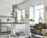 Nowoczesny salon z aneksem kuchennym, jasne wnętrze z białymi meblami, przezroczystymi krzesłami i szarą kanapą.