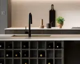 Nowoczesna kuchnia z wyspą, na której znajduje się blat slim line, czarny zlew i bateria. Wbudowane szafki na wino i stos talerzy.