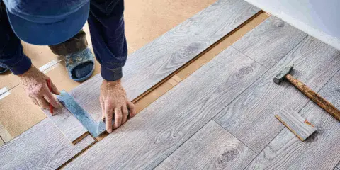 Ręczne docinanie paneli winylowych przy użyciu kątownika i ołówka. Obok leży młotek i przycięty fragment panelu.