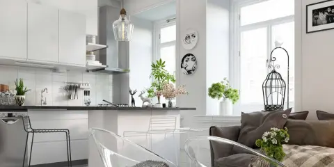 Nowoczesny salon z aneksem kuchennym, jasne wnętrze z białymi meblami, przezroczystymi krzesłami i szarą kanapą.
