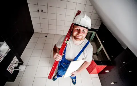 Budowlaniec w kasku i ogrodniczkach trzyma poziomicę i płytkę. Pokazuje, jak skuć płytki bez kurzu.