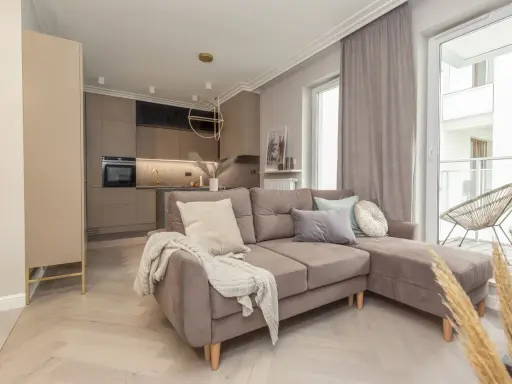 Nowoczesny salon z aneksem kuchennym. Dominują barwy monochromatyczne przykłady: beże, szarości, brązy. Miękka sofa, jasne drewno.