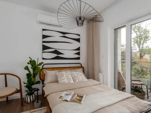 Przytulne lozko pod oknem z widokiem na balkon i zieleń. Wygodne poduszki i ciekawa grafika na ścianie tworzą relaksującą atmosferę.