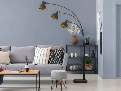 Sofa w kolorze grafitowym i szarym, ozdobiona poduszkami. Obok stolik kawowy i pufka.