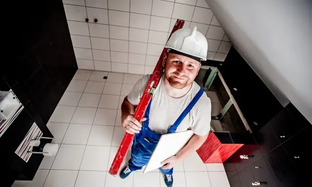 Budowlaniec w kasku i ogrodniczkach trzyma poziomicę i płytkę. Pokazuje, jak skuć płytki bez kurzu.