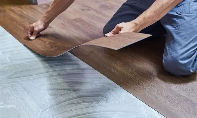 Ręce układają panel winylowy imitujący drewno na przygotowanym podłożu. Trwa wymiana panela winylowego.