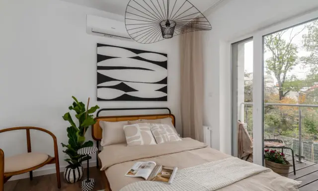 Przytulne lozko pod oknem z widokiem na balkon i zieleń. Wygodne poduszki i ciekawa grafika na ścianie tworzą relaksującą atmosferę.