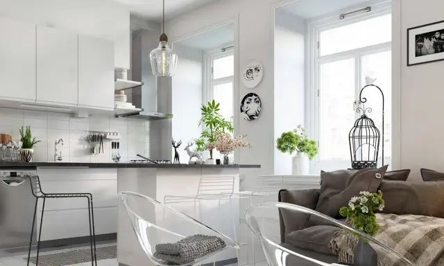Nowoczesny salon z aneksem kuchennym, jasne wnętrze z białymi meblami, przezroczystymi krzesłami i szarą kanapą.