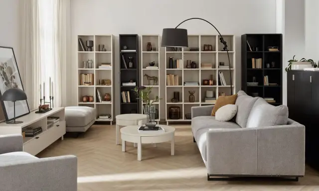 Nowoczesny salon beżowo szary z szarą kanapą, białymi regałami i czarną szafką.